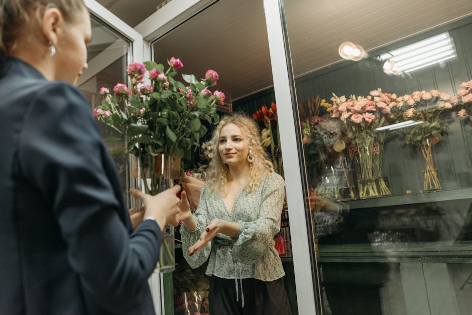 A florist at a flower shop, part of flowerswoking.co.uk, hands a large bouquet of pink roses with lush green foliage to a customer. The bouquet is wrapped in brown paper, and the flowers appear fresh 