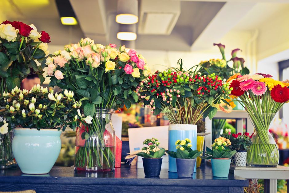 A variety of fresh floral arrangements displayed on a wooden table in a well-lit flower shop. The assortment includes bouquets of roses in shades of pink, white, and cream, arranged in clear glass and