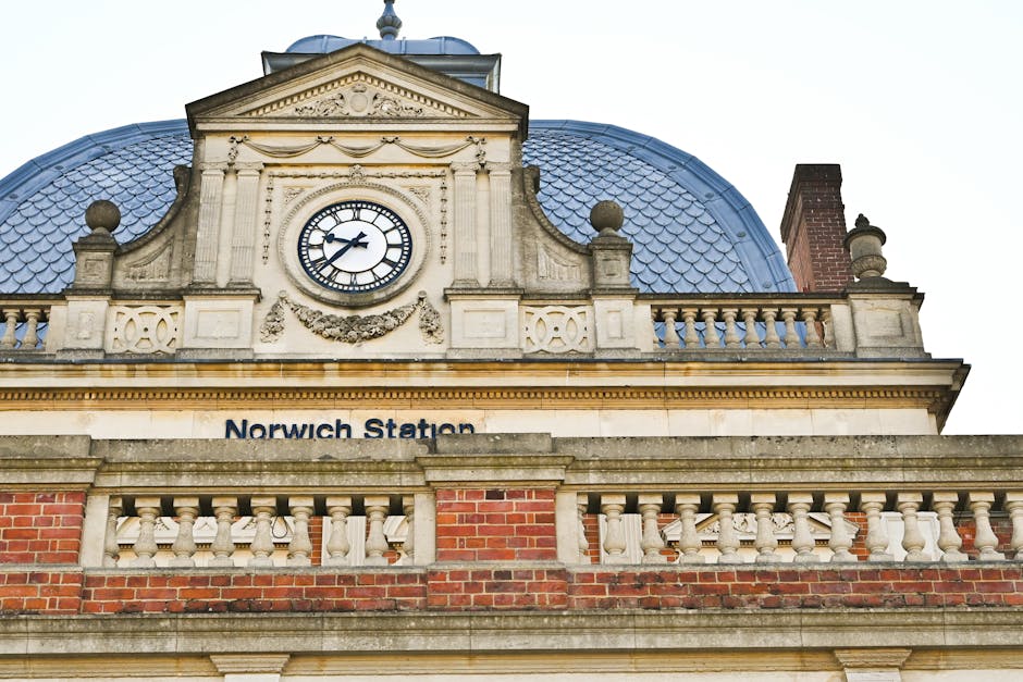 Close-up view of the upper section of Norwich Station's historic building showcasing a large clock with black Roman numerals and hands, set within an ornate stone façade above the station's sign. The